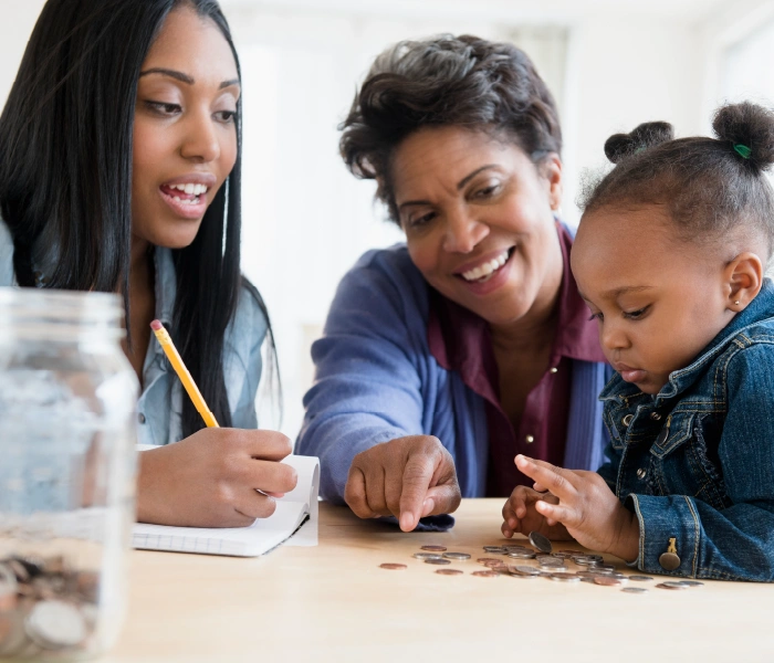 Three people at a table sorting coins. A woman writes on a notepad, another woman points at coins, and a child looks at them. A jar of coins is visible on the left.