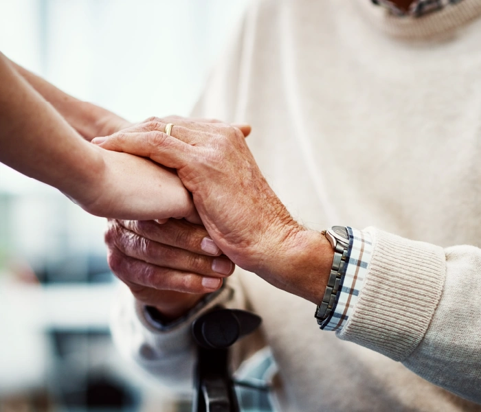 An elderly person holds someone's hand with both hands; the person wears a watch and a sweater.