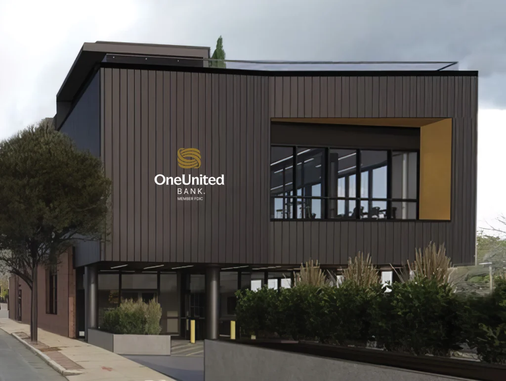 Modern bank building with "OneUnited Bank" logo, featuring a dark facade, large windows, and surrounding greenery.