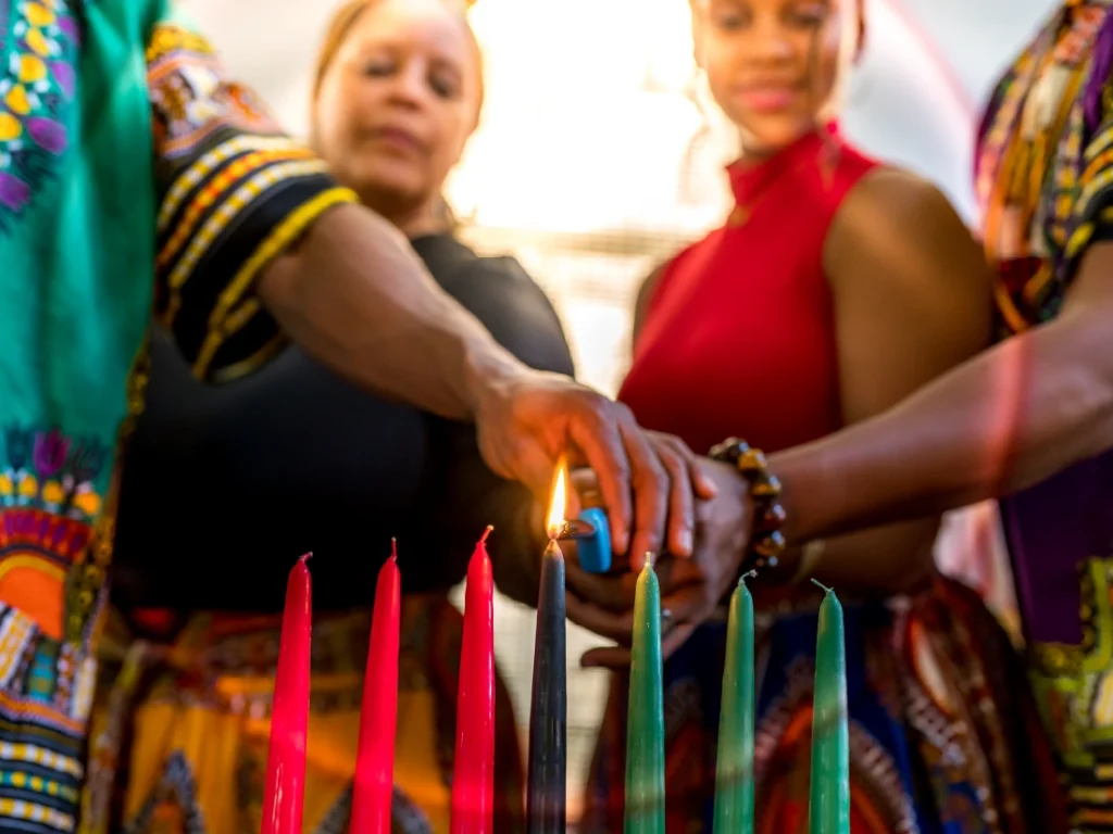 Three people dressed in colorful attire are lighting seven candles of different colors, possibly for a cultural or festive event like Kwanzaa.