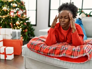 A woman in a red sweater lies on a couch, touching her temples with closed eyes. A Christmas tree and wrapped gifts are in the background.