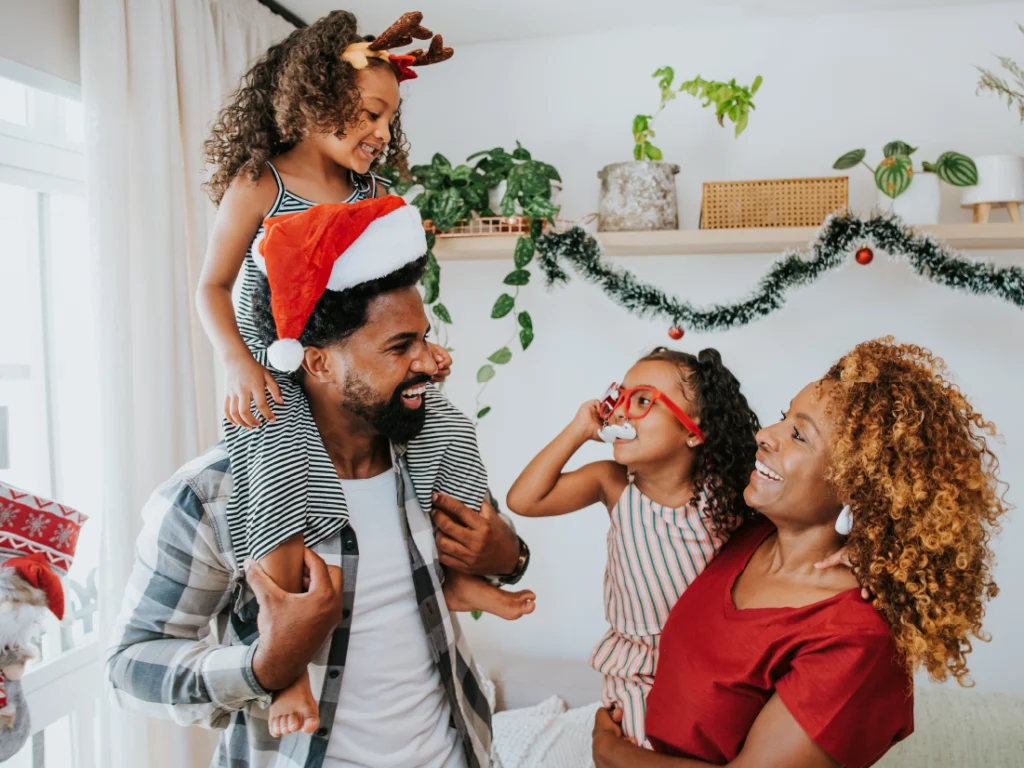 A family of four celebrates indoors. A man wears a Santa hat, carrying a child with reindeer antlers. A woman and another child with red glasses smile nearby. Holiday decor is visible.