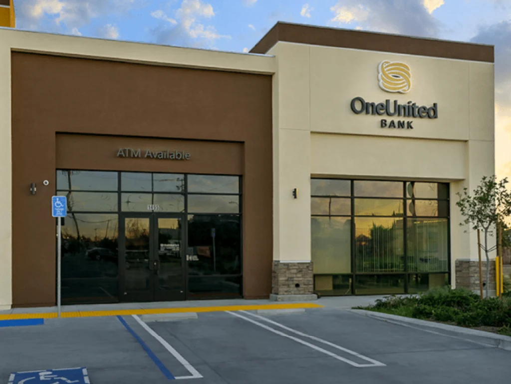 Exterior of OneUnited Bank with large windows and an ATM available sign. Accessible parking spot and trees are visible in the foreground.