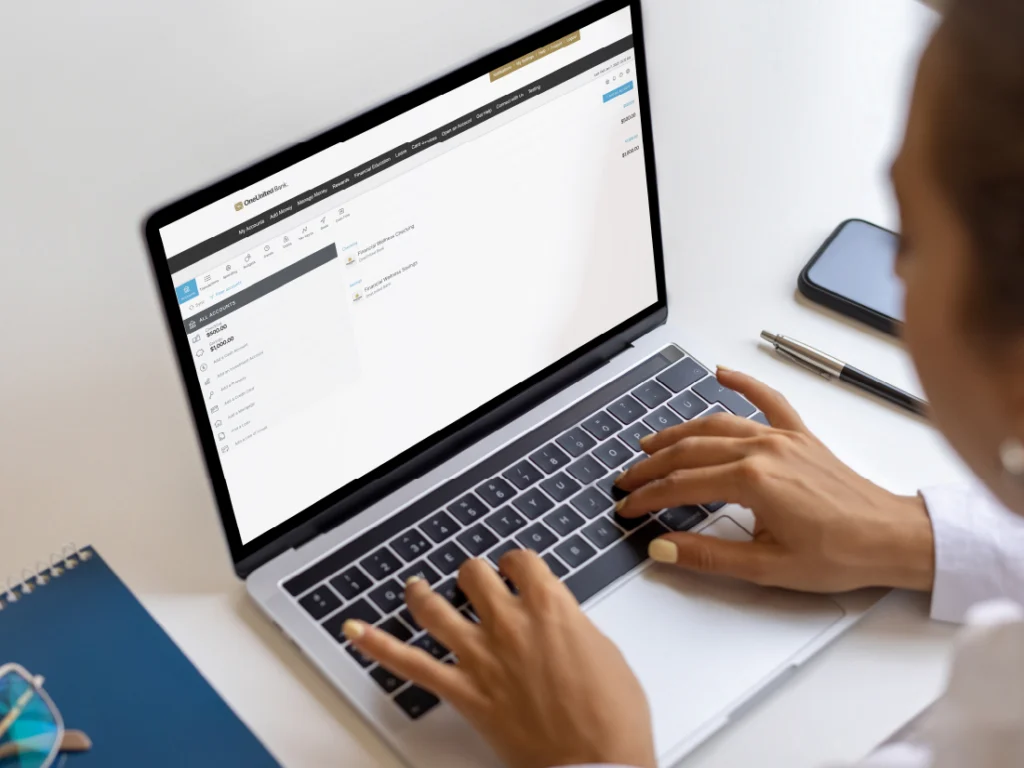 Person typing on a laptop with a website dashboard on the screen. A smartphone, pen, and notebook are on the desk.