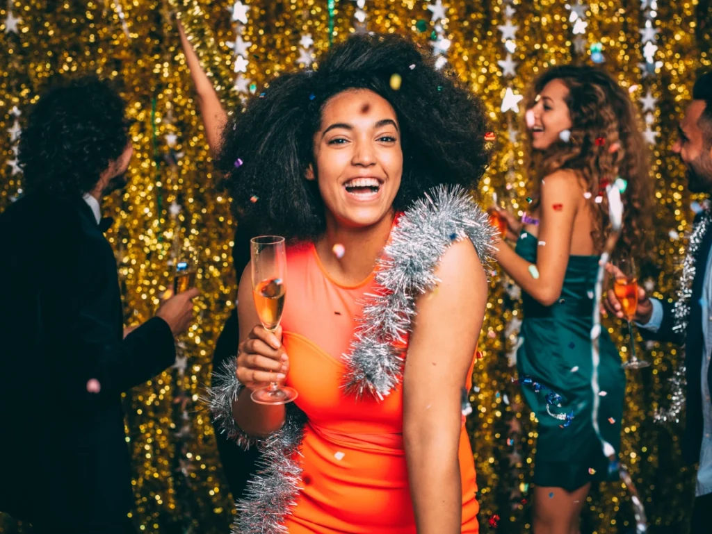 Woman in an orange dress holding a champagne glass, smiling at a party with a gold tinsel backdrop and people dancing in the background.