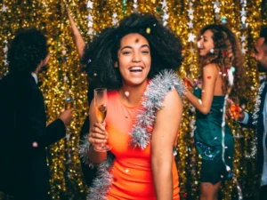 A woman in a red dress holds a champagne glass, smiling, with tinsel around her neck. People are celebrating in the background against a gold glitter backdrop.