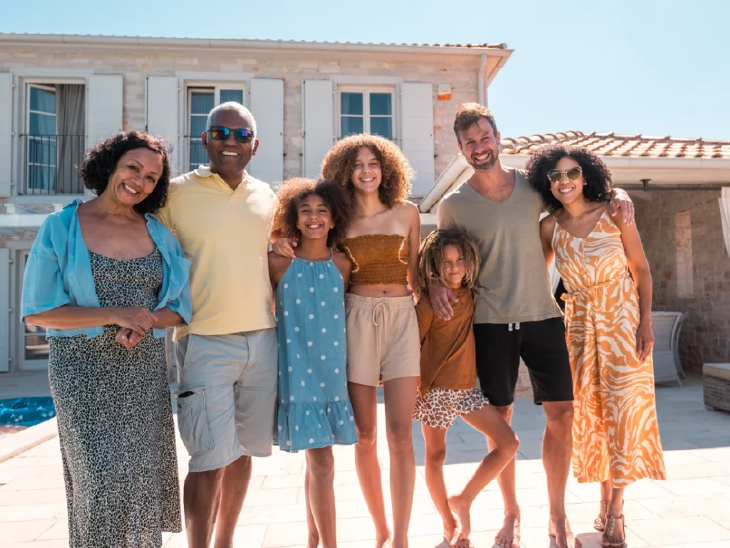 A group of seven people, including adults and children, stand smiling in front of their house with a pool. As proud new homeowners, they’re casually dressed and appear relaxed, enjoying the dream of homeownership.