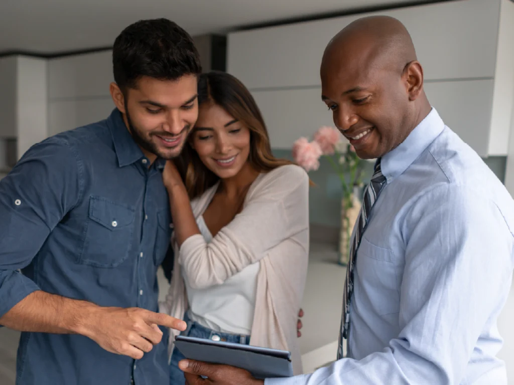 A couple looks at a tablet held by a man in a tie, standing in a modern kitchen, discussing their journey to homeownership.