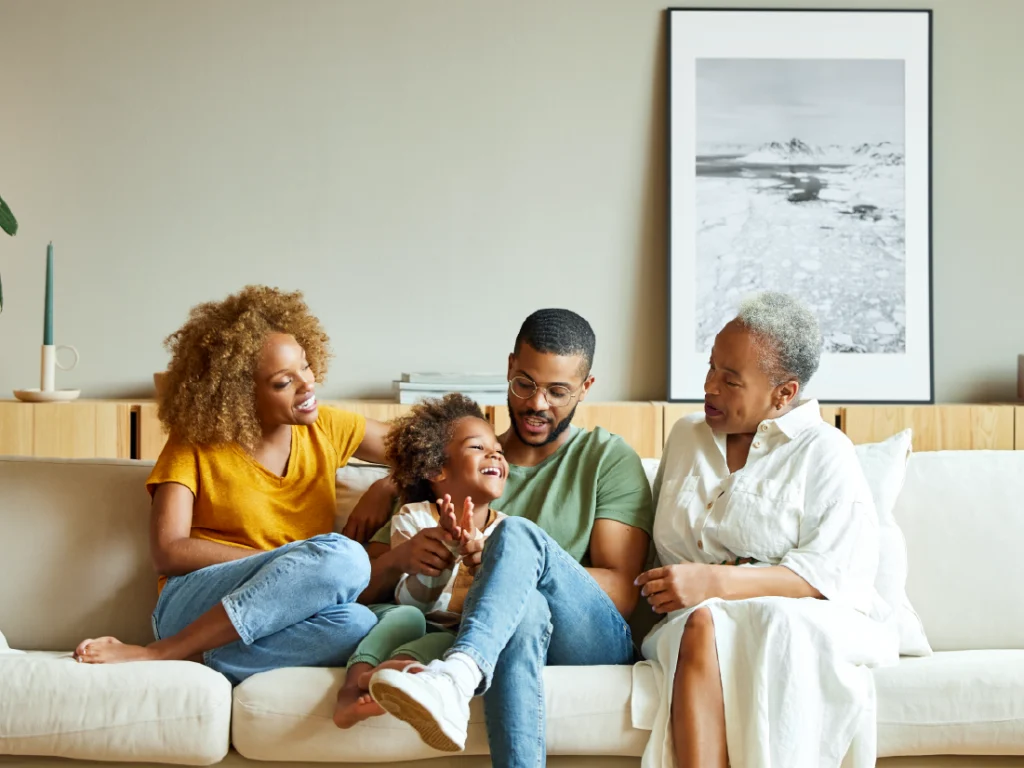 Four people, including a child, sit together on a sofa in their cozy living room, smiling and interacting. A black-and-white framed photo hangs on the wall, a testament to cherished memories and the joy of homeownership.