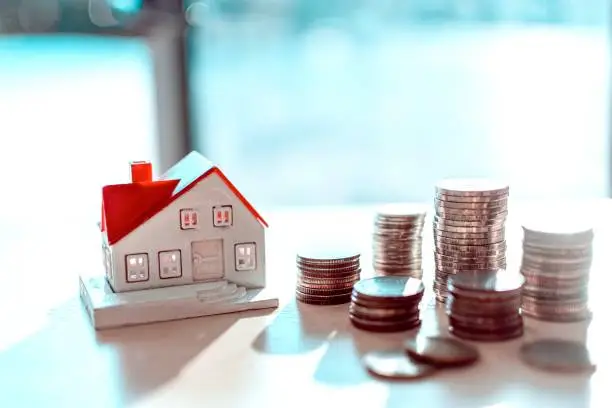 A small model house beside stacks of coins on a table, symbolizing real estate investment or savings.