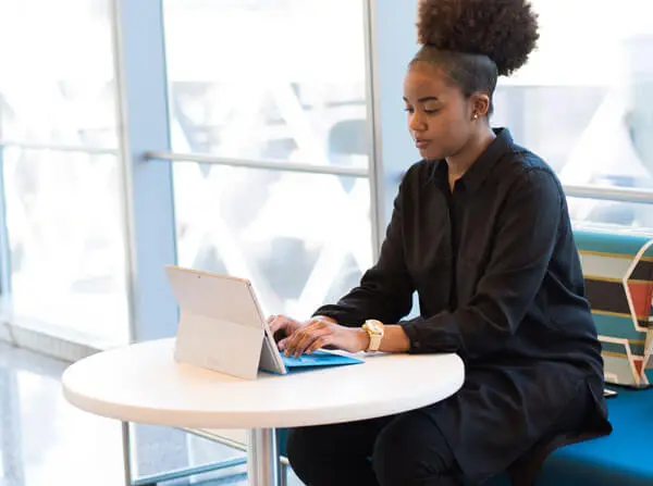 Person sitting at a round table, using a laptop. They're wearing a black outfit and a watch, with a colorful bag on the seat beside them. Bright window in the background.