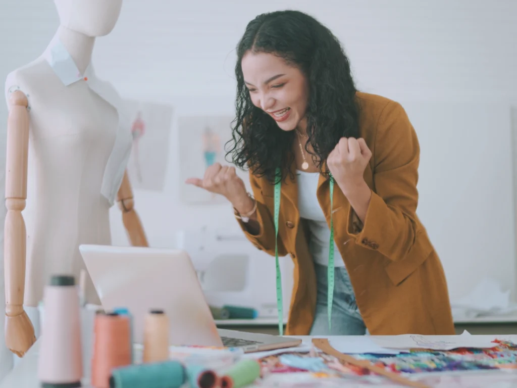 A woman in a mustard blazer and measuring tape smiles and clenches her fists at a laptop in her sewing studio, celebrating the success of her profitable business. A mannequin and colorful threads adorn the table, showcasing her vibrant creativity.