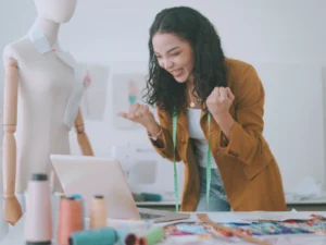 A woman in a mustard blazer and measuring tape smiles and clenches her fists at a laptop in her sewing studio, celebrating the success of her profitable business. A mannequin and colorful threads adorn the table, showcasing her vibrant creativity.