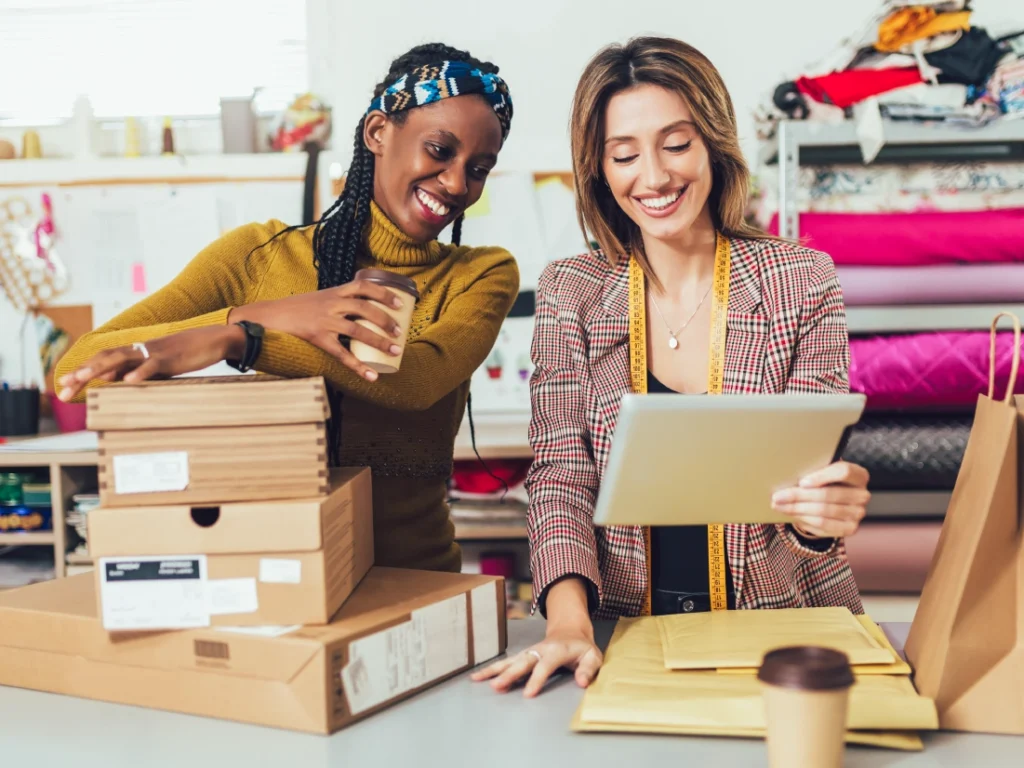 Two women in a workshop smile while handling packages and paperwork, creating the foundation for a profitable business. Various fabrics and a coffee cup rest on the table, symbols of their industrious collaboration.