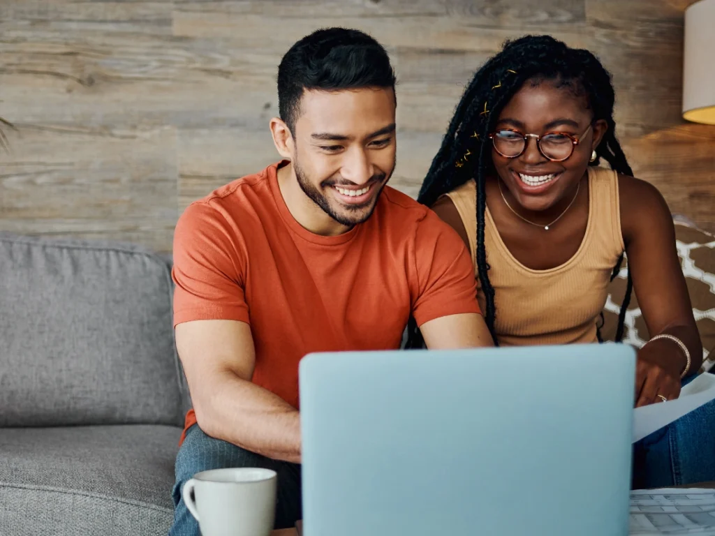 Two people, embracing the spirit of financial wellness, are smiling and looking at a laptop screen while sitting on a couch. A coffee cup is on the table in front of them.