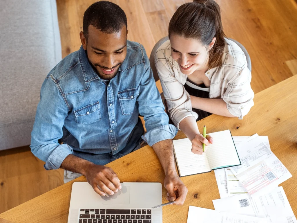 A man and woman are seated at a wooden table, laptop open among papers and a notebook, as they focus together on the screen, exploring paths to enhance their financial wellness.