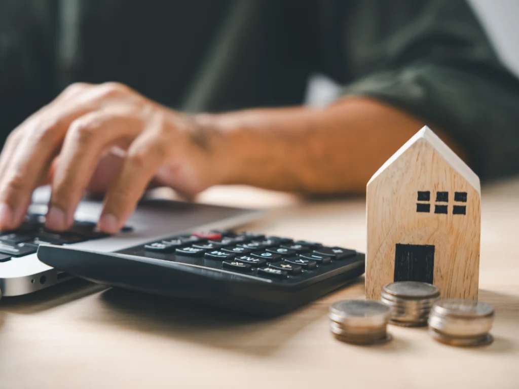 A person is typing on a laptop, considering refinancing options, with a calculator, miniature wooden house, and stacks of coins spread across the table.