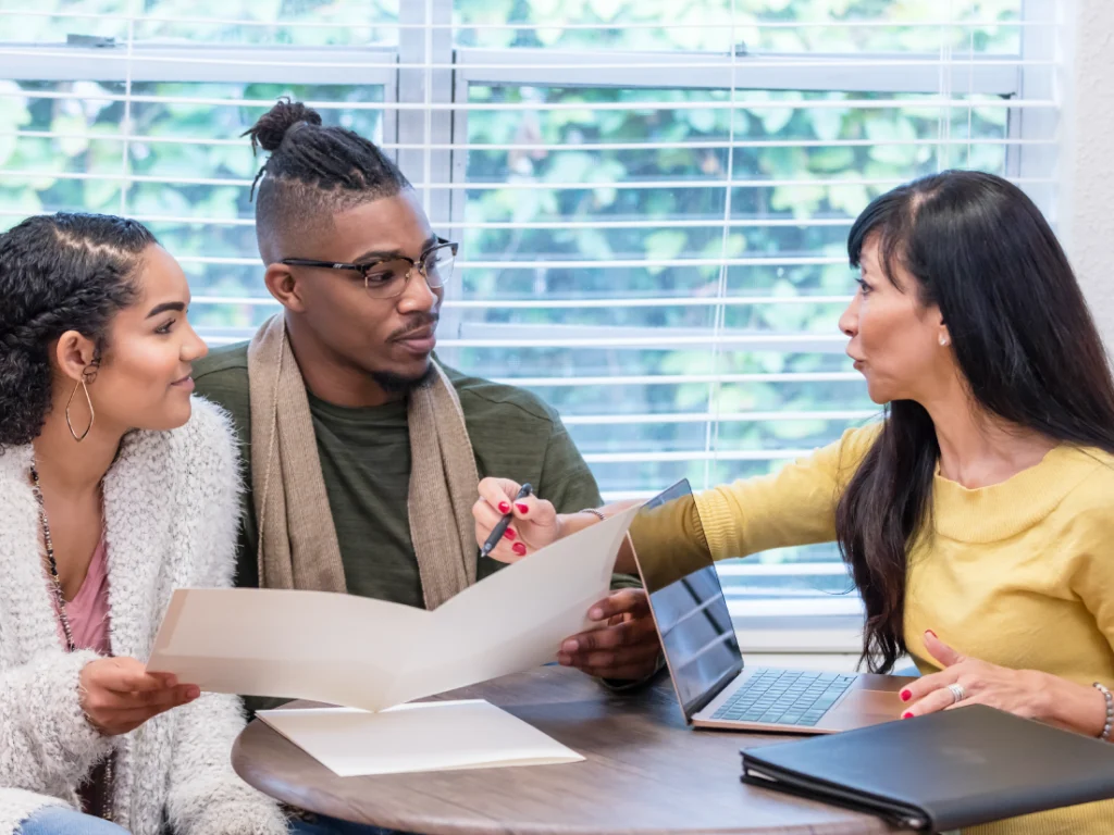 Three people sit at a table discussing refinancing documents. A woman on the right points at a folder held by a man in the center, while a woman on the left observes attentively.