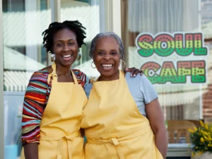 Two women in yellow aprons smile outside a cafe, proudly showcasing the "Soul Cafe" sign on the window of their family-owned business.