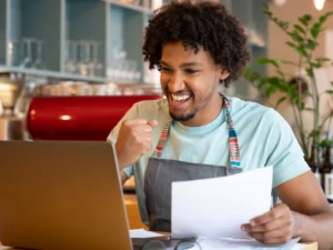 A person wearing an apron sits in front of a laptop, holding a document and smiling with a fist raised in triumph, likely celebrating success despite having an irregular income.