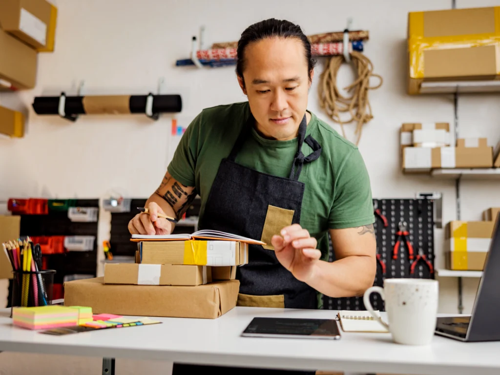 A person in an apron manages irregular income at a desk, with a notebook, packages, and a cup nearby, surrounded by storage items and tools.