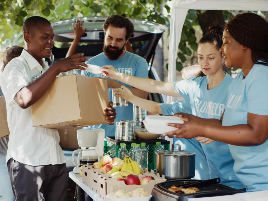 Volunteers in blue shirts distribute food and drinks to a man holding a cardboard box at an outdoor event, highlighting the community spirit. Not only does volunteering lend a helping hand, but it also comes with tax benefits for those generously giving their time.