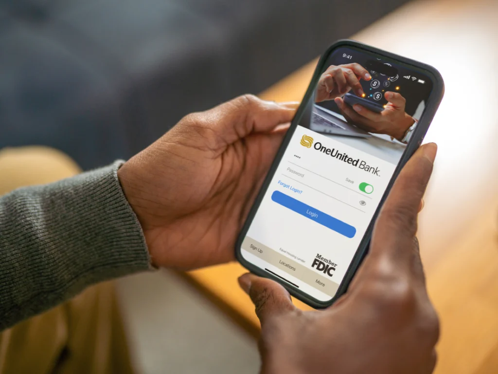 A person holds a smartphone showing the FDIC-insured OneUnited Bank login screen.